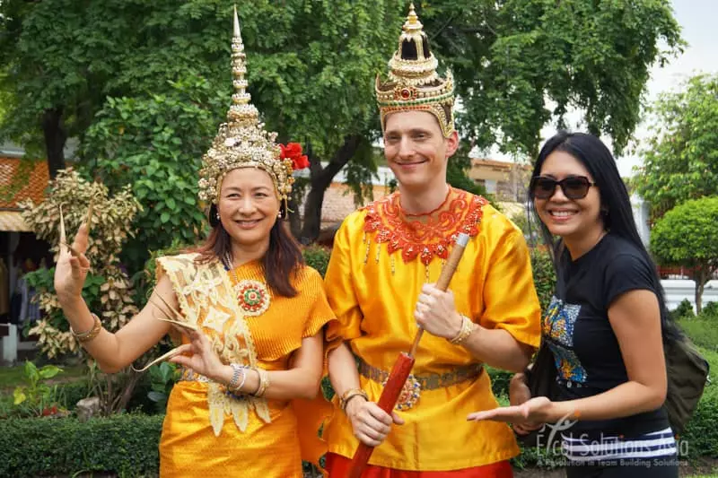 A team adorned in gold traditional Thai costume, standing in awe before the majestic Wat Arun temple in Bangkok during the Amazing Race.