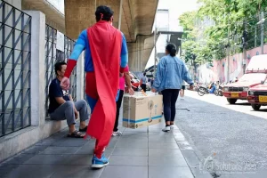 Superhero team demonstrating kindness and teamwork at Saphan Taksin, Bangkok, Thailand, by assisting a lady with a heavy parcel during a team-building event.